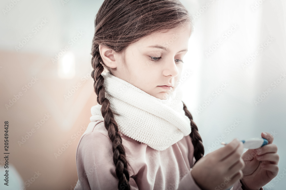 Have a cold. Serious little female covering neck with scarf and bowing head while looking at thermometer Have a cold. Serious little female covering neck with scarf and bowing head while looking at thermometer
