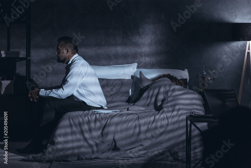 Dark gray room with a side view of a mature man sitting on a bed and having second thoughts after cheating and a woman sleeping under the cover photo