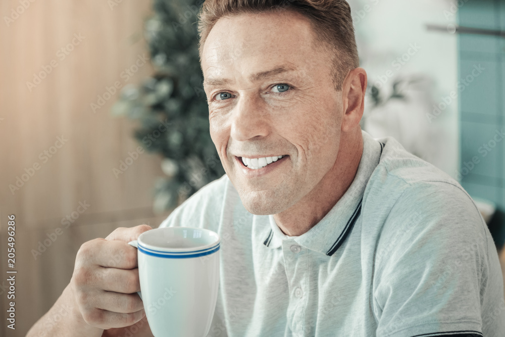 Time at home. Attractive male person keeping smile on his face and holding cup in right hand while drinking tea Time at home. Attractive male person keeping smile on his face and holding cup in right hand while drinking tea