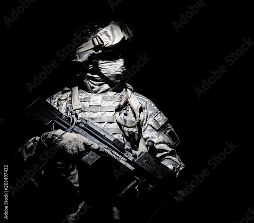 Billede på lærred United States Armed Forces soldier in battledress with black glasses and mask on face, armed squad automatic weapon emerges from darkness