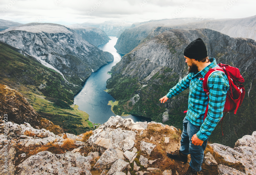 Naklejka premium Travel Man with backpack standing on mountain top exploring Norway lifestyle adventure hiking vacations aerial view Naeroyfjord success wellness healthy life concept