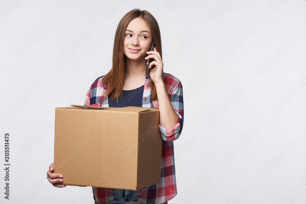 Delivery, relocation and unpacking. Smiling young woman holding cardboard box and talking on cellphone looking away at blank copy space. Call center and customer support service concept. Delivery, relocation and unpacking. Smiling young woman holding cardboard box and talking on cellphone looking away at blank copy space. Call center and customer support service concept.