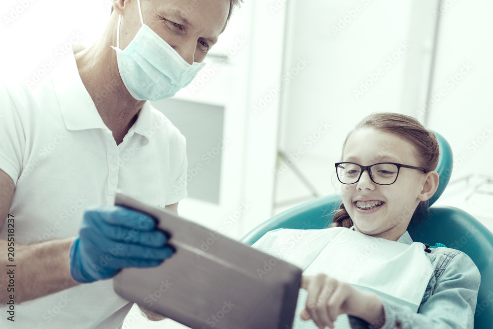 So interesting. Smiling little girl looking at the screen of a tablet while sitting in a dental chair with a professional doctor by her side So interesting. Smiling little girl looking at the screen of a tablet while sitting in a dental chair with a professional doctor by her side