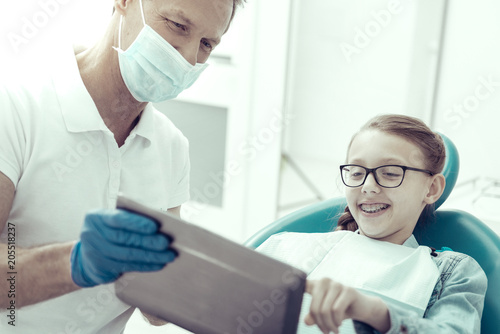 So interesting. Smiling little girl looking at the screen of a tablet while sitting in a dental chair with a professional doctor by her side