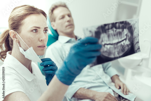 Small changes. Responsible serious young dentist looking attentively at the X-ray while her patient sitting in a dental chair