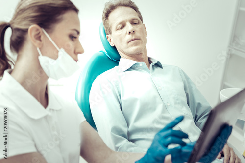 Detailed explanation. Serious concentrated patient listening to the recommendations of his dentist while sitting in a dental chair and looking at the screen of a tablet