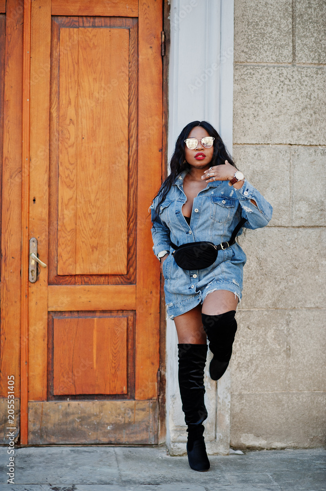 African american girl in jeans dress and sunglasses posed on streets of city. Black stylish model shoot.