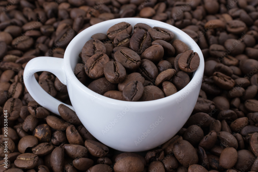 White cup full of coffee beans on the background of coffee beans. Close up White cup full of coffee beans on the background of coffee beans. Close up