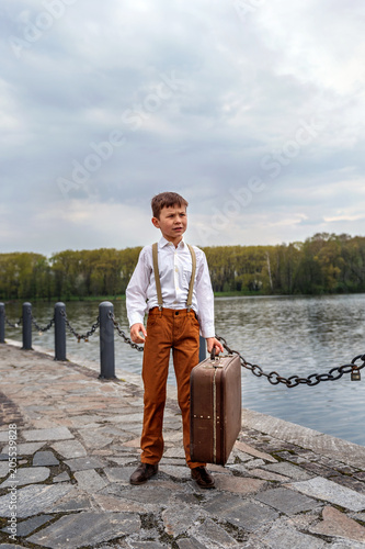 little boy simpleton in an old-fashioned rustic clothes with a vintage suitcase standing at the arrival station on the pier of the lake river