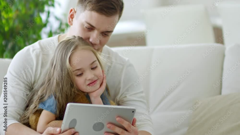 Medium shot of cute little girl sitting on fathers laps and watching cartoons on tablet Medium shot of cute little girl sitting on fathers laps and watching cartoons on tablet