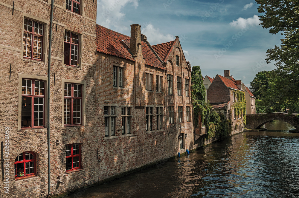 Wooded gardens and brick buildings on the canal's edge in a sunny day at Bruges. With many canals and old buildings, this graceful town is a World Heritage Site of Unesco. Northwestern Belgium.