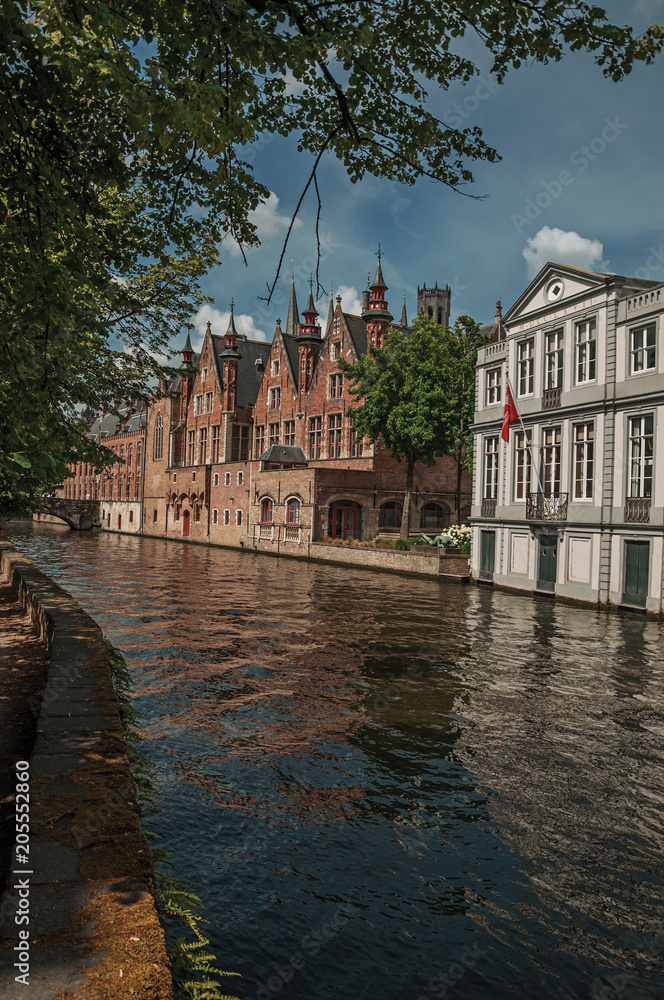 Obraz premium Wooded gardens and brick buildings on the canal's edge in a sunny day at Bruges. With many canals and old buildings, this graceful town is a World Heritage Site of Unesco. Northwestern Belgium.