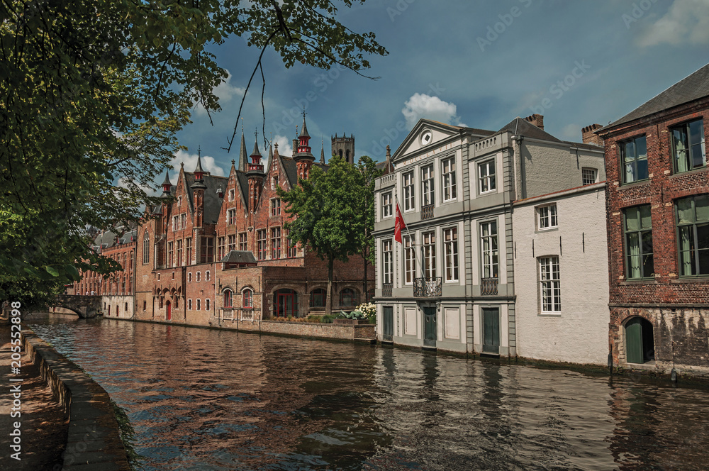 Obraz premium Wooded gardens and brick buildings on the canal's edge in a sunny day at Bruges. With many canals and old buildings, this graceful town is a World Heritage Site of Unesco. Northwestern Belgium.