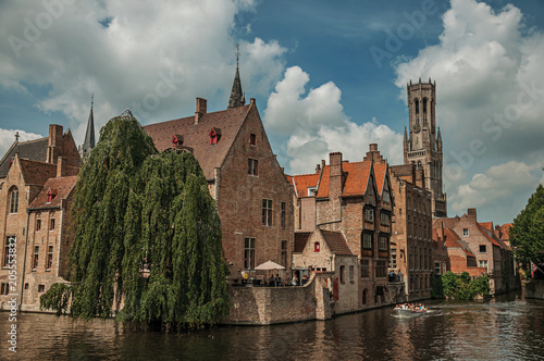 Boats and leafy tree with brick buildings on the canal's edge in a sunny day at Bruges. With many canals and old buildings, this graceful town is a World Heritage Site of Unesco. Northwestern Belgium.