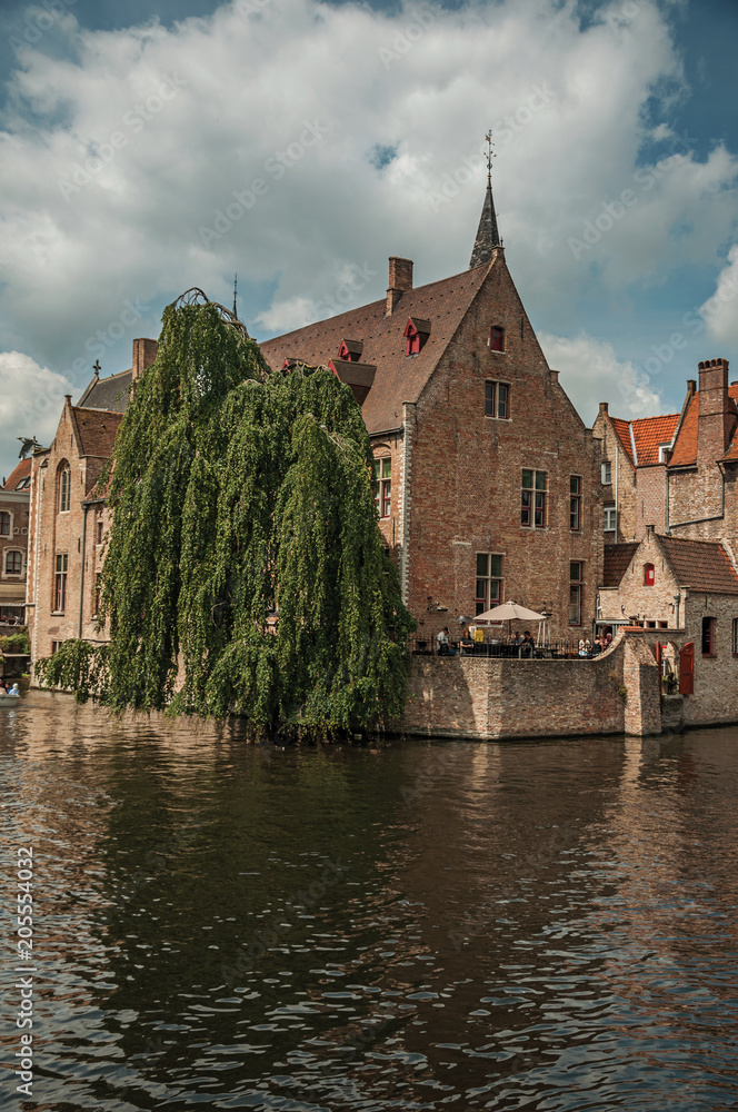 Leafy tree with old brick buildings on the canal's edge in a sunny day at Bruges. With many canals and old buildings, this graceful town is a World Heritage Site of Unesco. Northwestern Belgium. Leafy tree with old brick buildings on the canal's edge in a sunny day at Bruges. With many canals and old buildings, this graceful town is a World Heritage Site of Unesco. Northwestern Belgium.