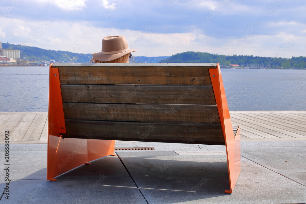 Drewniana ławka z czerwonymi metalowymi bokami, od tyłu, na betonowo-drewnianym nabrzeżu, jakby molo, na niej siedzi turysta w słomkowym kapeluszu i ciemnych okularach, patrzy na zatokę i drugi brzeg - obrazy, fototapety, plakaty 