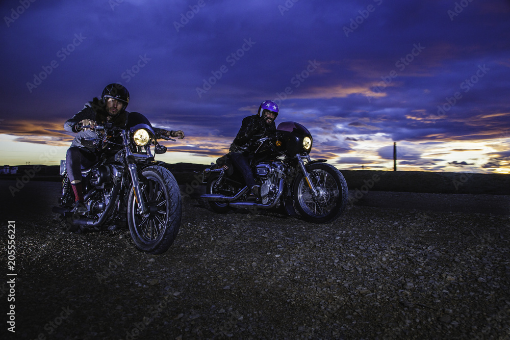 Naklejka premium Modern bikers ride on a closed road with helmets on classic motorcycle at sunset. Outdoor portrait and urban lifestyle