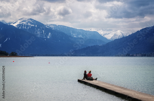 Young woman and young man sitting together on the jetty in the tegernsee lake and looking at the blue mountains in the famous tourist resort of the Bavarian Alps, Bavaria, Germany, Europe,