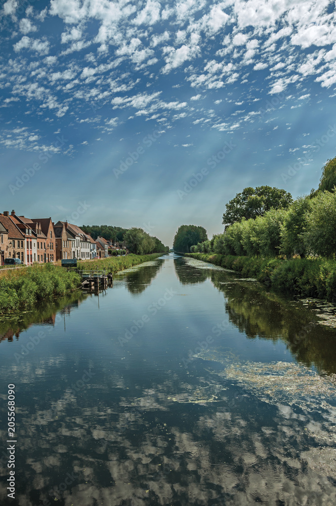 Houses, bushes and grove along canal with sky reflected on water, in the late afternoon and blue sky, near Damme. A quiet and charming countryside old village near Bruges. Northwestern Belgium. Houses, bushes and grove along canal with sky reflected on water, in the late afternoon and blue sky, near Damme. A quiet and charming countryside old village near Bruges. Northwestern Belgium.