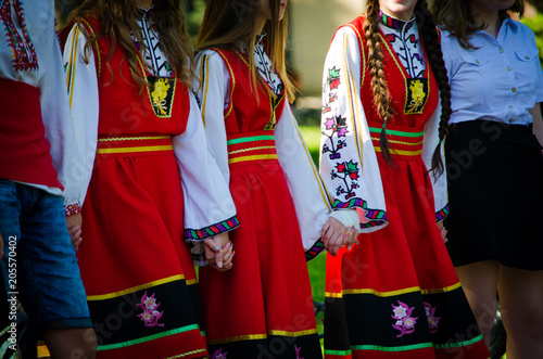Unrecognizable girls in traditional Bulgarian costumes with red dresses and patterns on white shirts holding hands