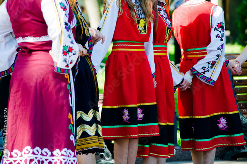 Unrecognizable girls in traditional Bulgarian costumes with red dresses and patterns on white shirts holding hands
