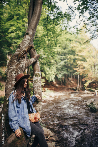 stylish traveler girl in hat sitting on tree and looking at river in sunny forest. summer vacation. travel and wanderlust concept. space for text. woman traveling in mountains