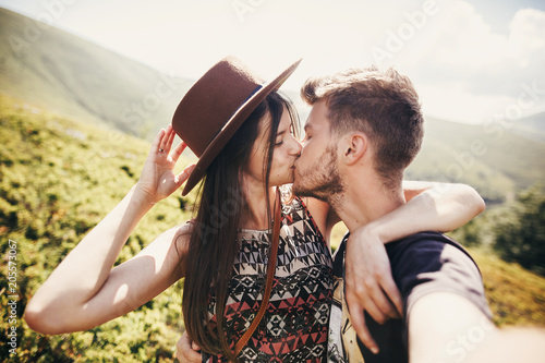 stylish hipster couple taking selfie and kissing on top of mountains in sunny summer day. beautiful man and happy woman in hat embracing on honeymoon. travel together photo