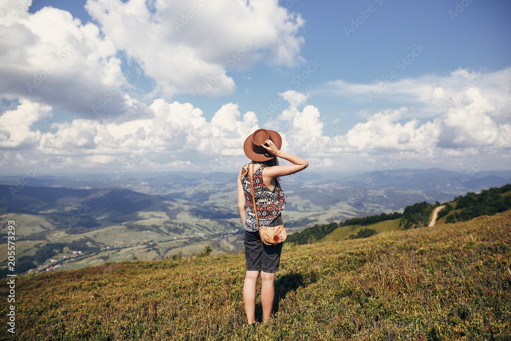 Fototapeta premium travel and wanderlust concept. stylish traveler girl in hat looking at sunny mountains and clouds sky. summer vacation. space for text. back view. amazing atmospheric moment