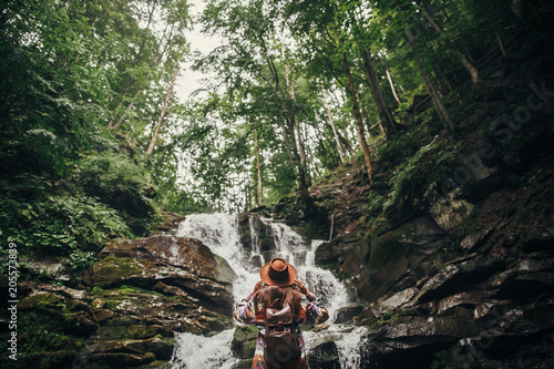 stylish hipster girl in hat with backpack looking at waterfall in forest in mountains. traveler woman exploring woods. travel and wanderlust concept. atmospheric moment