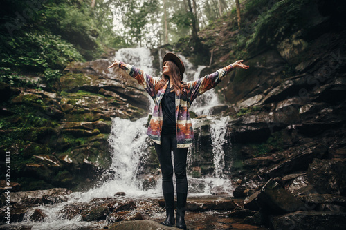 travel and wanderlust concept. stylish hipster girl in hat with backpack standing at waterfall in forest in mountains. traveler woman raising hands up in woods. atmospheric moment