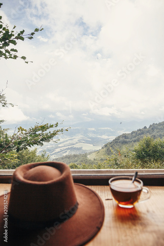 hipster hat and tea in glass on wooden table at window light with view on mountains and sky. delicious hot drink on mountain top. summer travel and wanderlust. focus on mountain