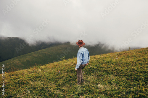 stylish traveler man in hat walking on top of sunny mountains into the  misty clouds. travel and wanderlust concept. space for text. happy hipster guy traveling. summer vacation © sonyachny