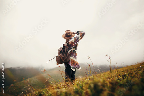 travel and wanderlust concept. stylish traveler hipster girl holding hat, with backpack and windy hair, walking in mountains in clouds. summer vacation. space for text. amazing atmospheric moment