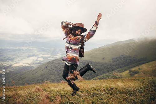 stylish traveler hipster girl in hat, jumping with backpack and smiling in mountains in clouds. space for text. amazing atmospheric funny moment. travel and wanderlust concept