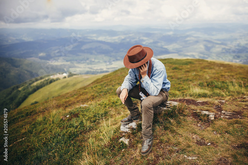 stylish traveler man sitting on top of mountains in clouds. space for text. hipster guy traveling, talking on phone. amazing atmospheric moment. travel and wanderlust concept