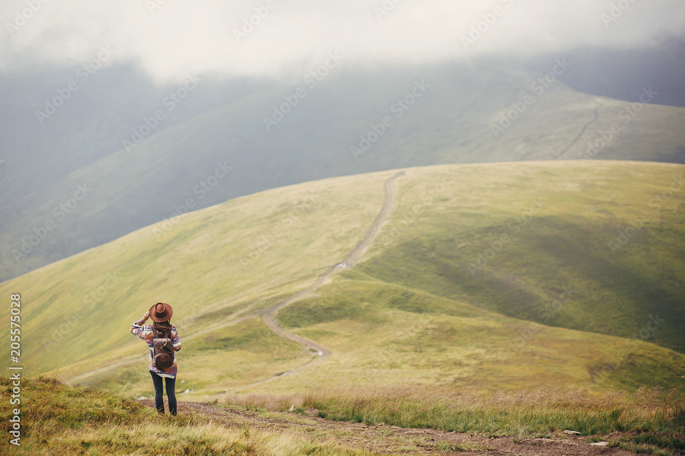 Fototapeta premium travel and wanderlust concept. traveler hipster girl in hat with backpack exploring misty mountains, hill with path. space for text. stylish woman traveling. amazing atmospheric moment.