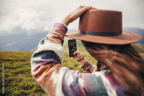 stylish traveler girl in hat with windy hair holding phone on top of sunny mountains. summer vacation. travel and wanderlust concept. space for text. back view. amazing atmospheric moment