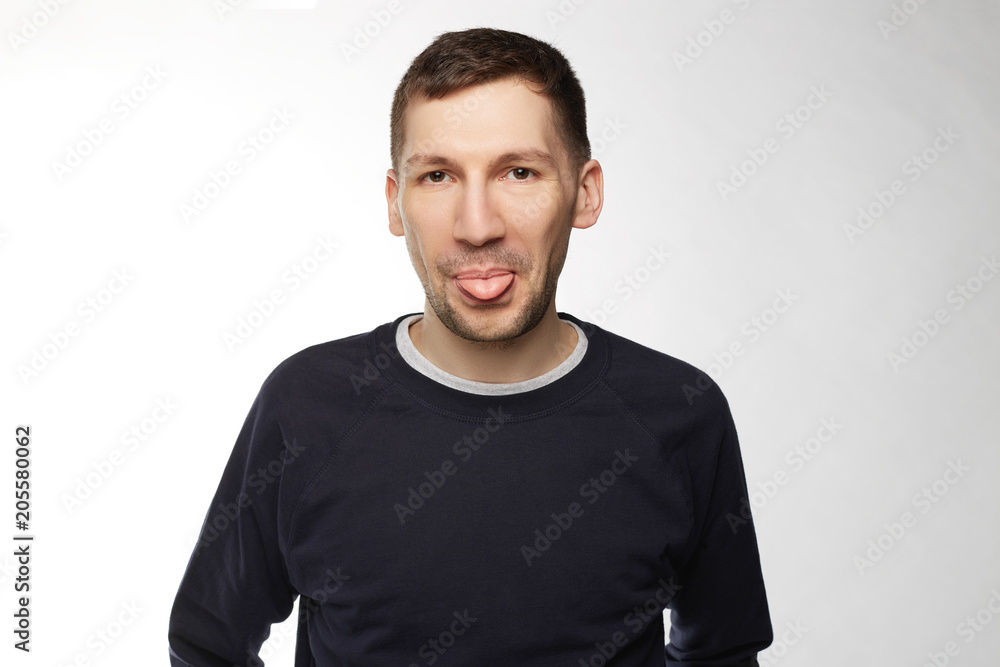 Portrait of handsome young male with brunette hair and pleasant appearance, shows tongue, has fun, wearing dark blue casual sweater, going to trick a friend. Health, Beauty and youth concept. Portrait of handsome young male with brunette hair and pleasant appearance, shows tongue, has fun, wearing dark blue casual sweater, going to trick a friend. Health, Beauty and youth concept.