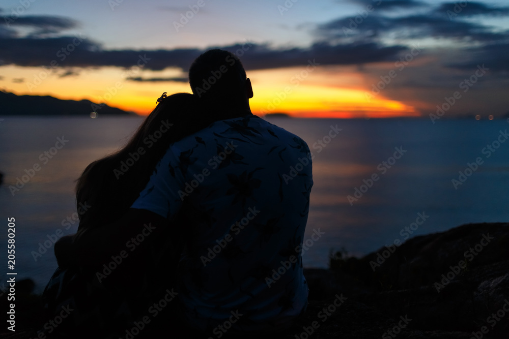 Back view Silhouette of a couple are sitting and hugging on the beach at sunset.