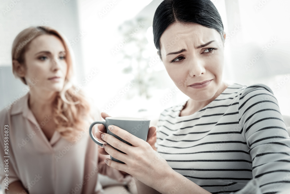 Dont cry. Unhappy sad disappointed woman sitting in the bright room on the sofa holding a cup and looking aside. Dont cry. Unhappy sad disappointed woman sitting in the bright room on the sofa holding a cup and looking aside.