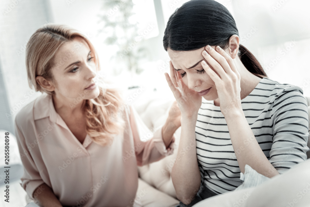 Bed feeling. Exhausted sad cute woman sitting near her friend on the sofa touching her head closing eyes. Bed feeling. Exhausted sad cute woman sitting near her friend on the sofa touching her head closing eyes.