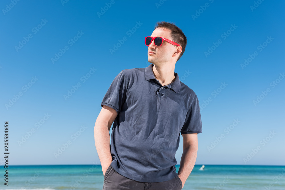 Young handsome attractive man hipster millennial serious face closeup on beach during sunny day with red sunglasses in Florida panhandle with ocean, hands in pockets