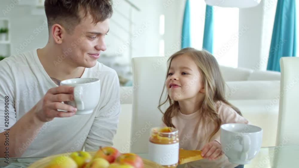 Medium shot of happy young father drinking tea and chatting with cute little daughter while having breakfast in morning Medium shot of happy young father drinking tea and chatting with cute little daughter while having breakfast in morning