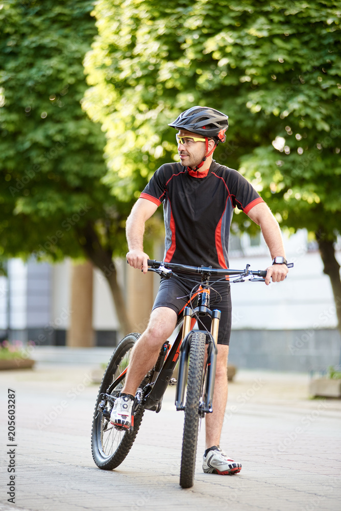 Fototapeta premium Guy professional cyclist wearing cycling clothes and helmet standing near bicycle looking to side interested and curious. Sportsman having break after hard training. Concept of healthy lifestyle
