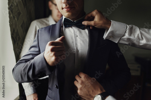 stylish groomsmen helping happy groom getting ready in the morning for wedding ceremony. luxury man in suit in room. space for text. wedding day. photo