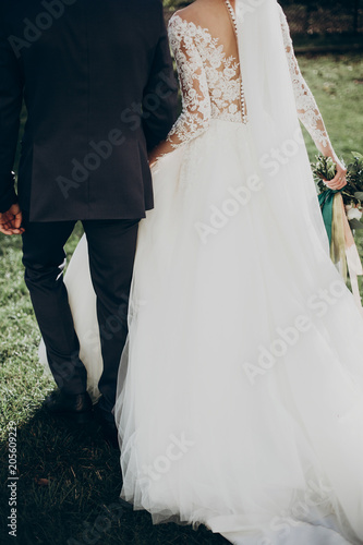 stylish happy bride and groom walking in yard and holding hands at wedding ceremony. emotional moment, space for text. luxury wedding newlyweds couple. back view