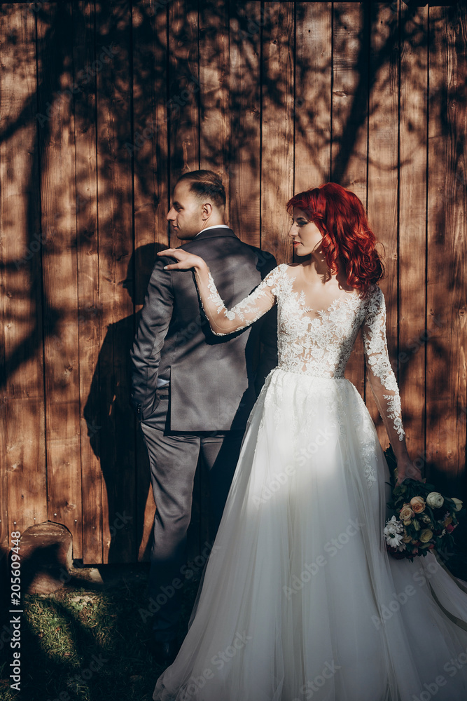 stylish bride and groom posing in sunlight on background of wooden wall in country. rustic wedding concept in barn, space for text. happy bohemian newlyweds, modern couple