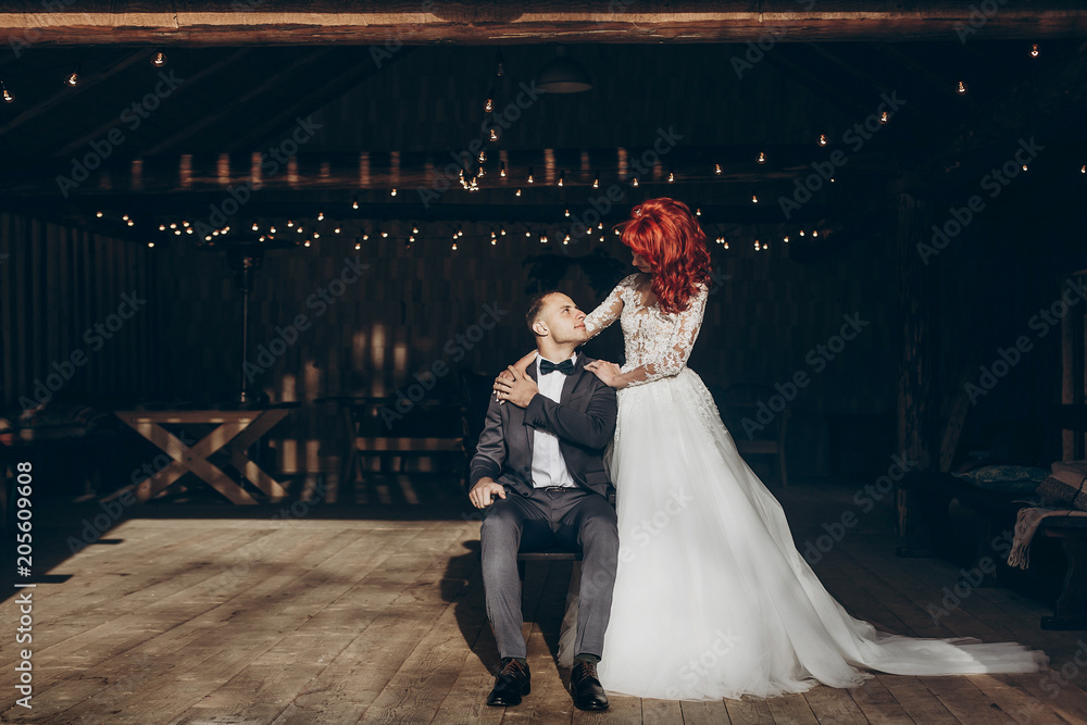 stylish groom sitting and happy bride hugging him on background of wooden barn with retro lights. rustic wedding concept, space for text. newlyweds couple embracing, romantic moment stylish groom sitting and happy bride hugging him on background of wooden barn with retro lights. rustic wedding concept, space for text. newlyweds couple embracing, romantic moment