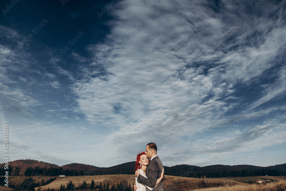 happy stylish bride and groom hugging in mountains in sunset light. gorgeous boho newlywed couple embracing, true feelings. emotional romantic moment. space for text, amazing view.
