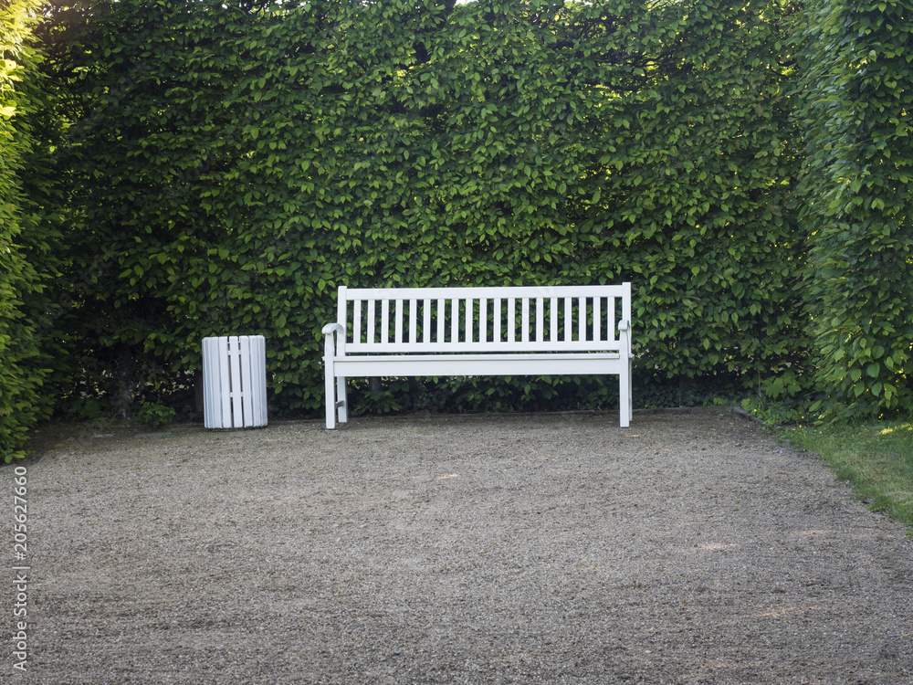 Ein weiße Bank aus Holz steht vor einer grünen Hecke in einem Park.